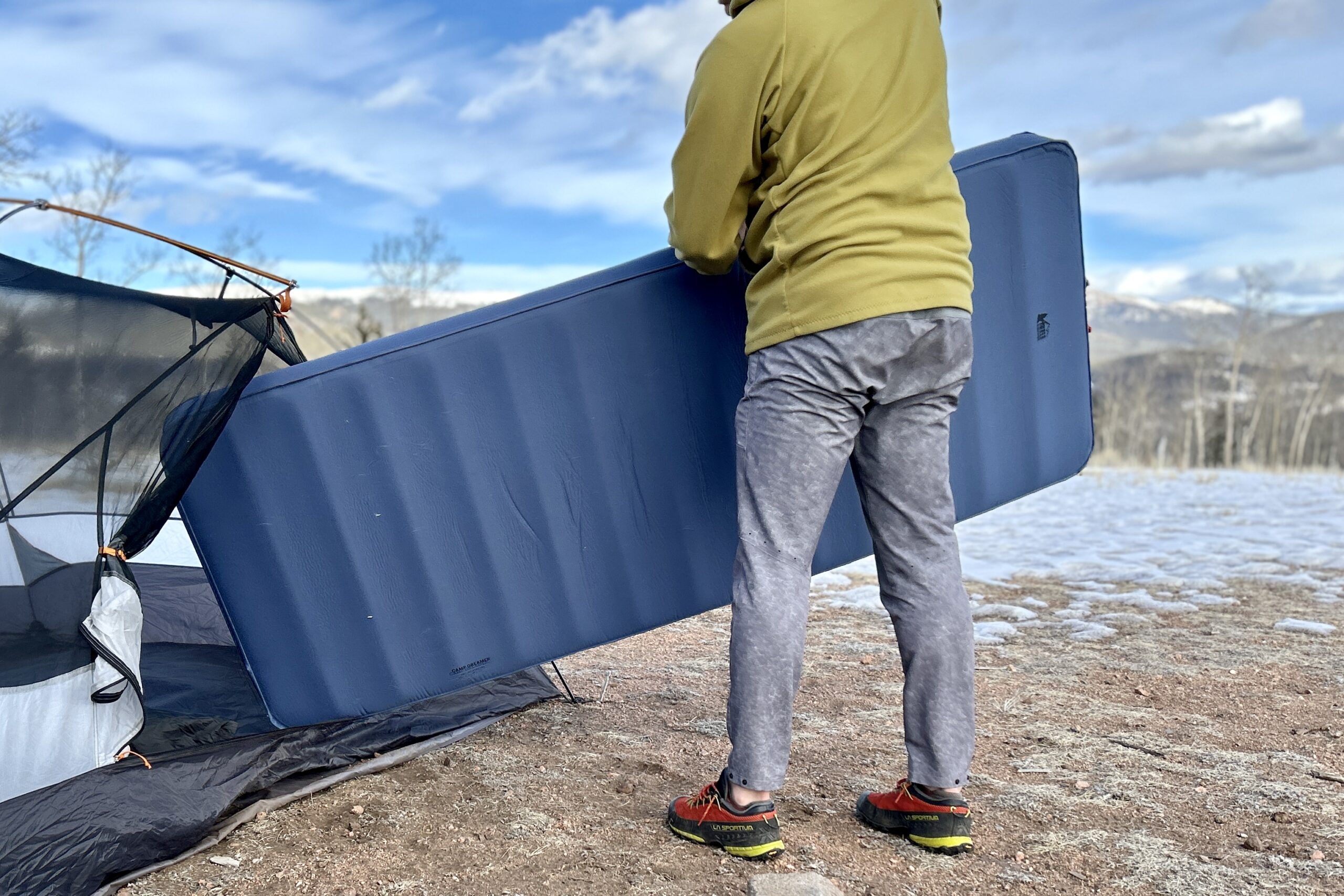 A man puts a sleeping mattress into a tent with no fly on. There is a snowy, mountainous landscape in the background.