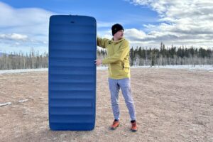 A man in a snowy, forested landscape stands next to a tall sleeping pad.