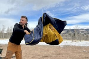 A man fluffs a sleeping bag outside in a snowy mountain landscape.