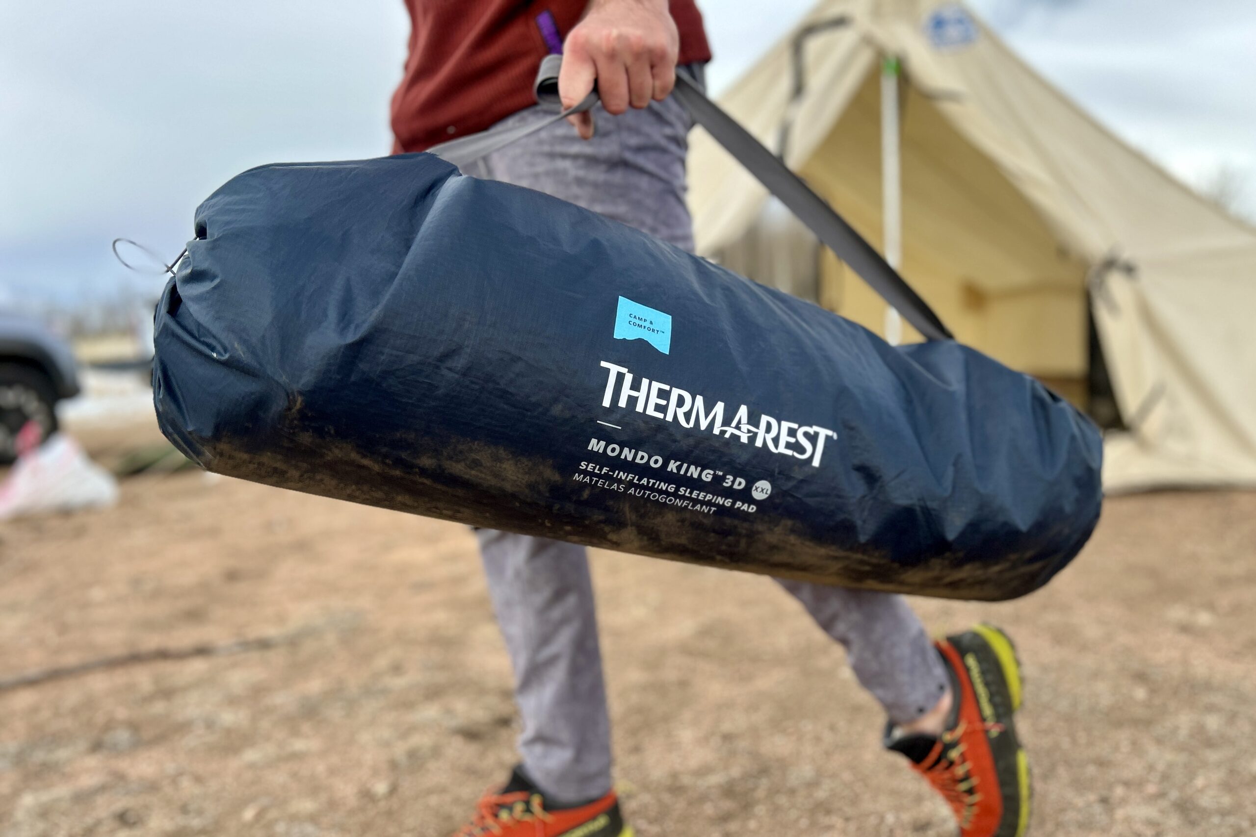 Close-up image of a person from the waist down carrying a packed-up sleeping mattress with it's thick nylon handle.