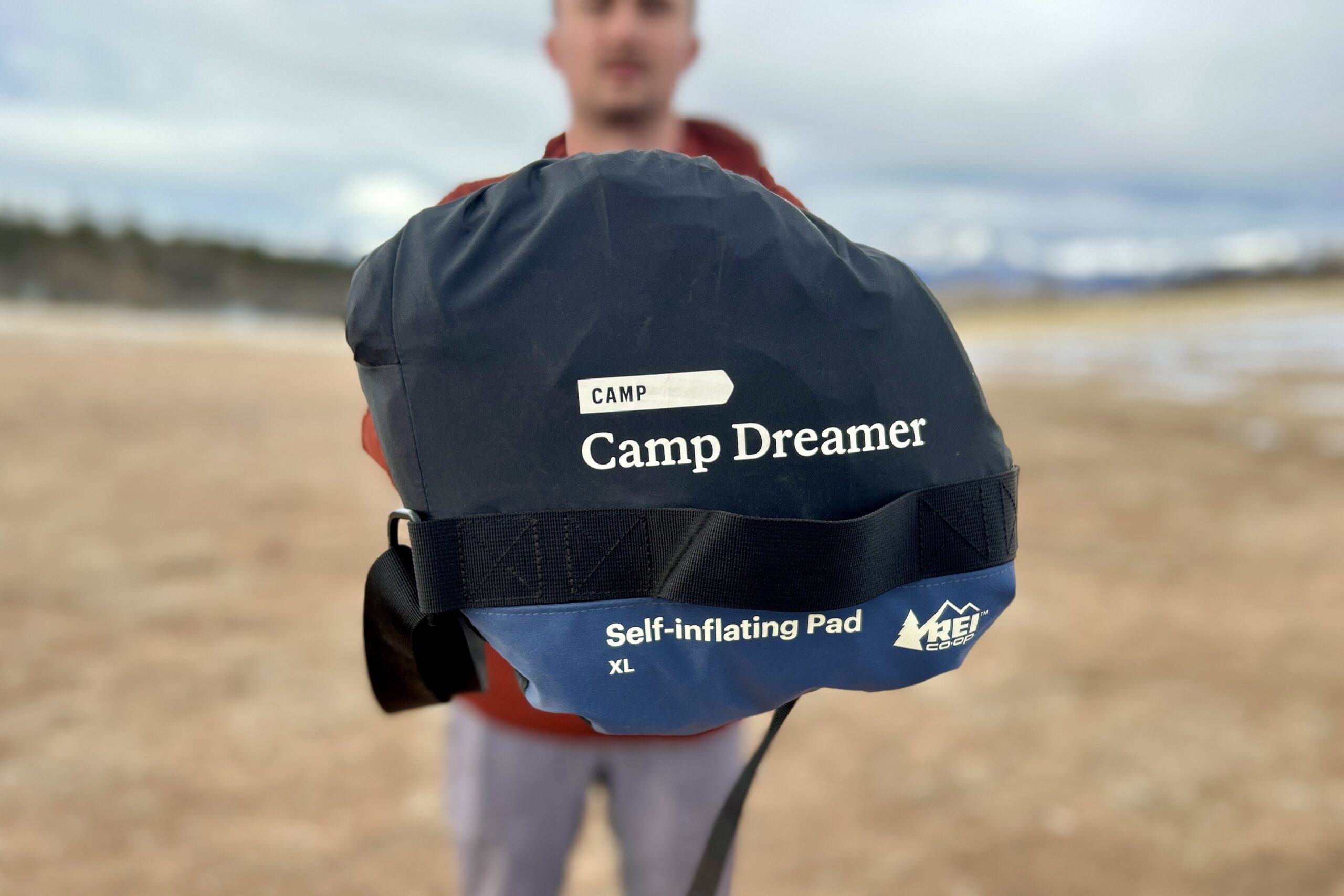 A person holds up a sleeping pad in a package to show off the bottom, where the text reads "CAMP DREAMER Self-inflating Pad XL" with the REI CO-OP logo.