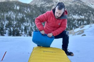 A man in a red jacket rolls up a pump sack attached to a camping sleeping pad in a wintry setting.