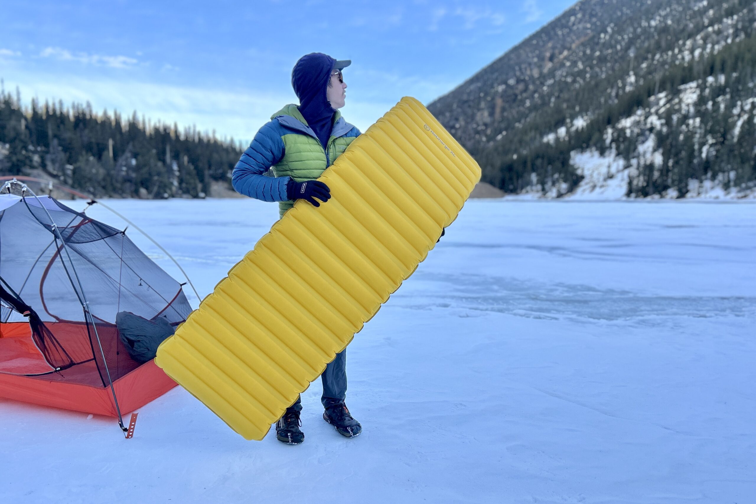 A man holds onto a camping sleeping pad outside a tent in a wintry setting.