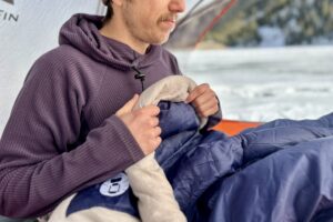 Close up image of a man sitting up in a sleeping bag in a tent in a wintry setting with their hands on the small draft tube at the collar.