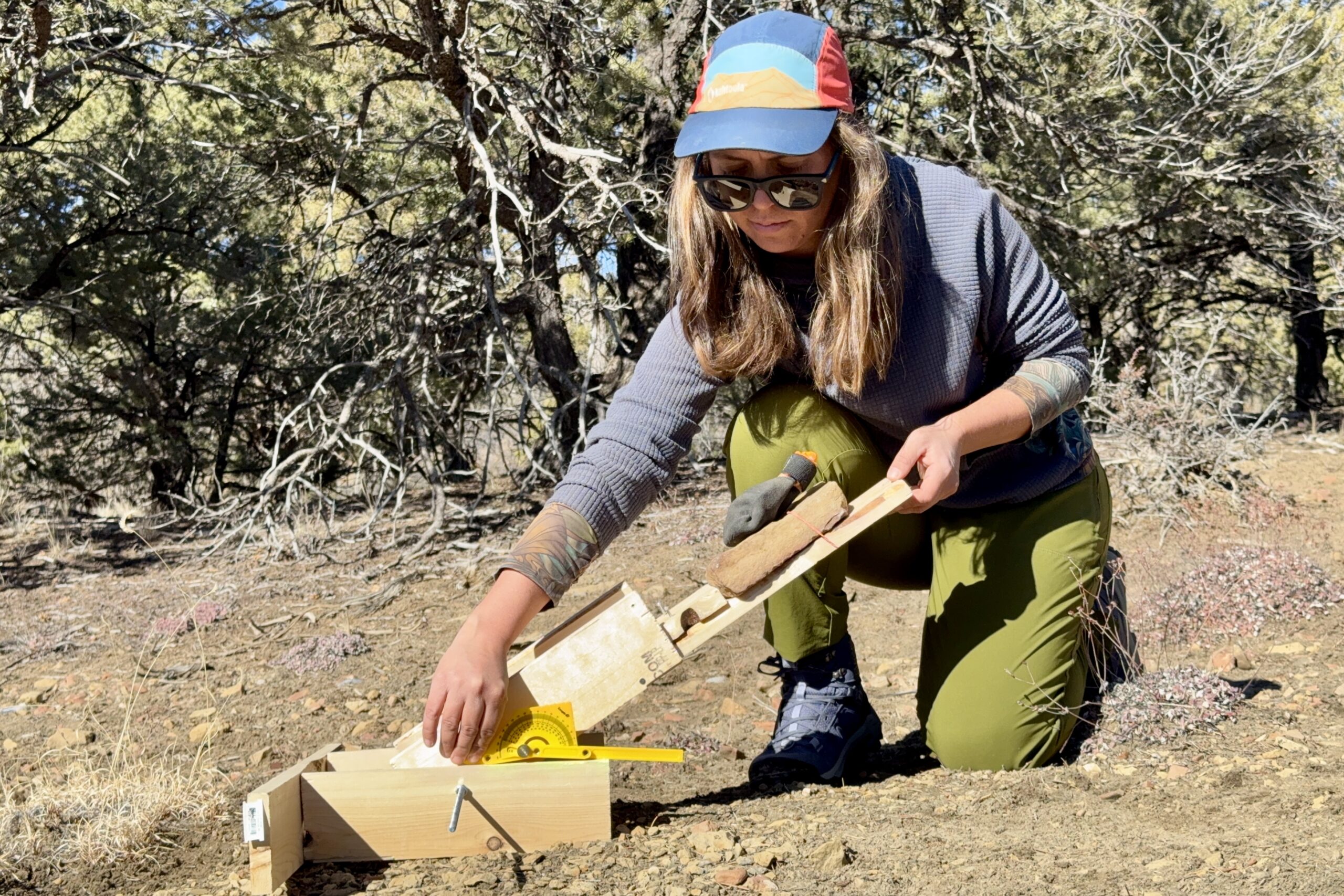 A person is testing boot traction on the Protector boots. They are holding up a wooden board with a rock on top. The boot is on top of the rock. They are also holding a protractor to measure the angle of the board. The test is being performed in a pinion forest.