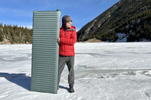 A man holds a camping mattress vertically in a snowy mountain setting.