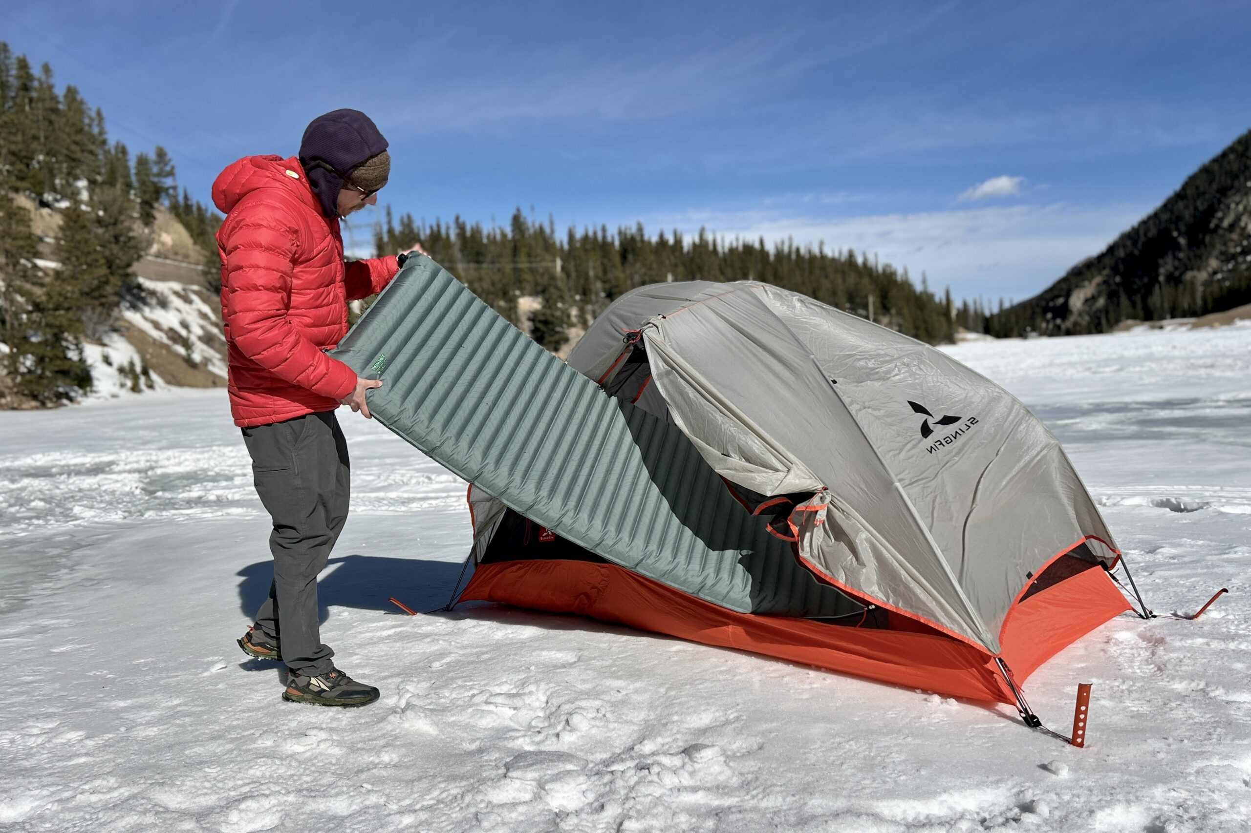 A man slides a camping mattress into a tent in a wintry setting.