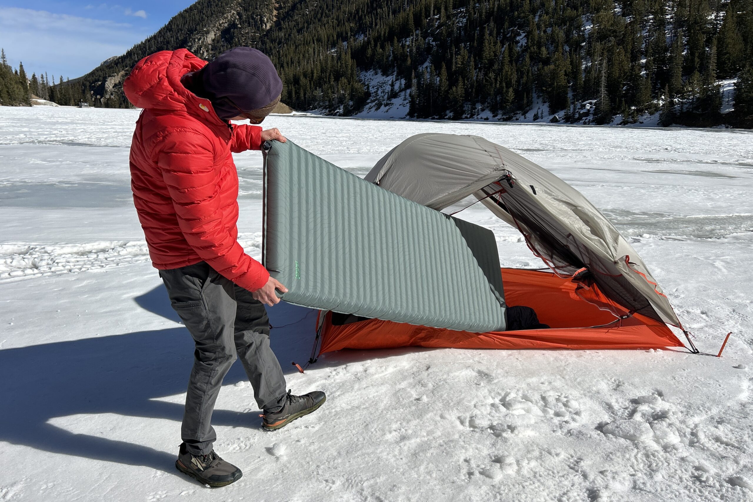A person slides a camping mattress into a tent in a wintry setting.