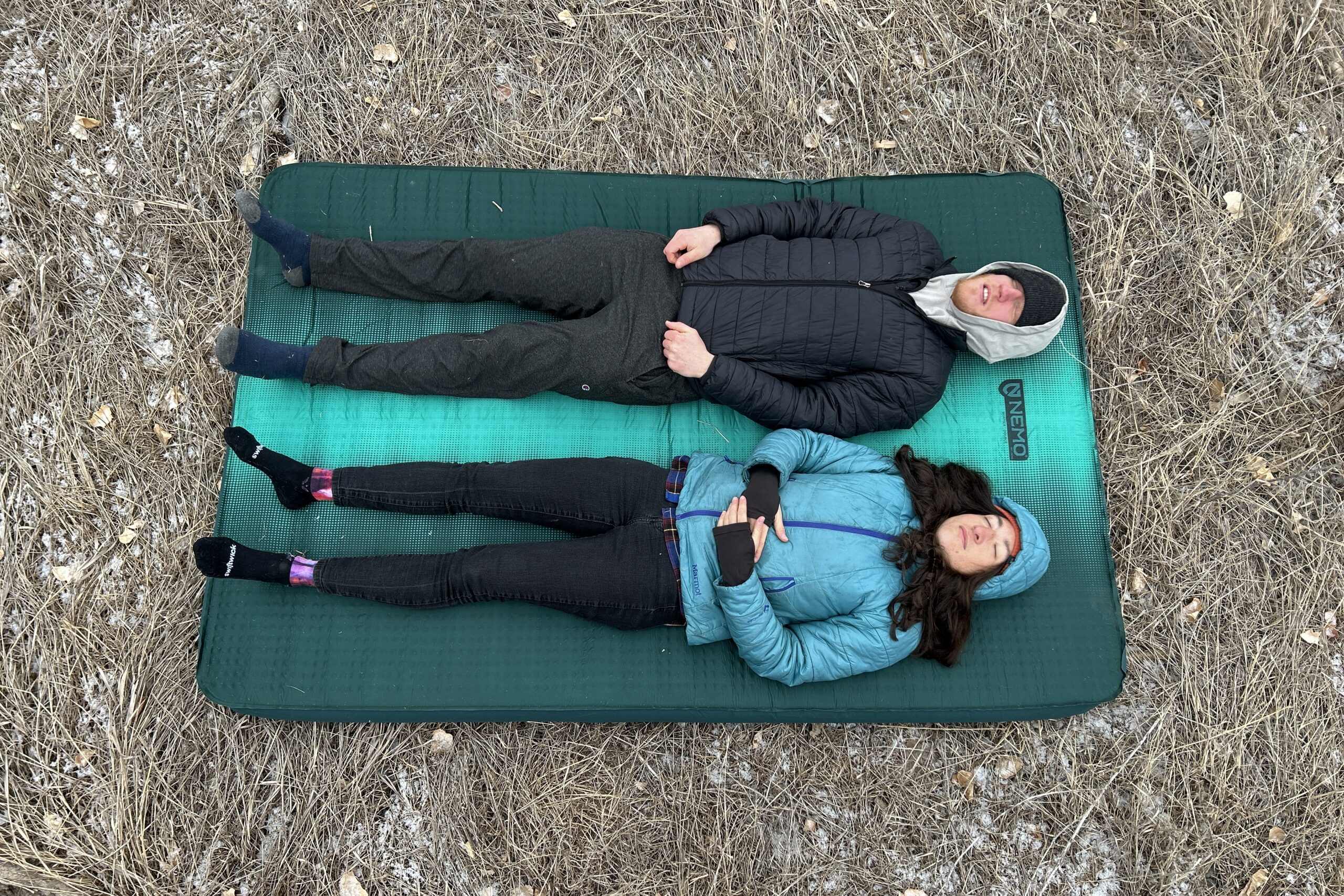 Top-down image of two people resting on a camping mattress on the grass.