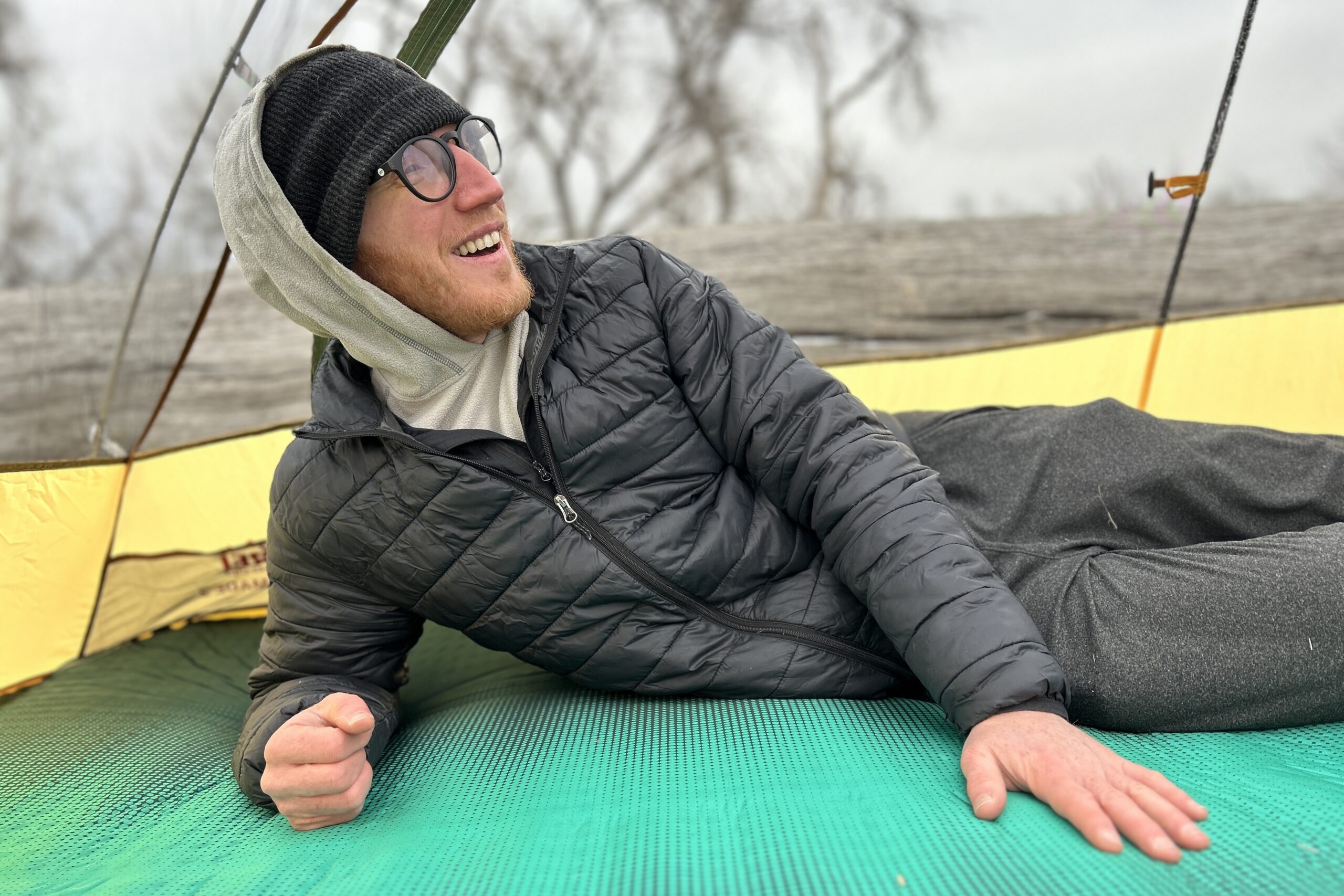 Close-up image of a man laying on a camping mattress inside a tent. He is smiling and looking off in the distance.