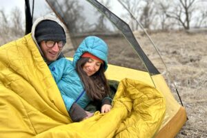 A man and woman are smiling and laying in a double sleeping bag in a tent.