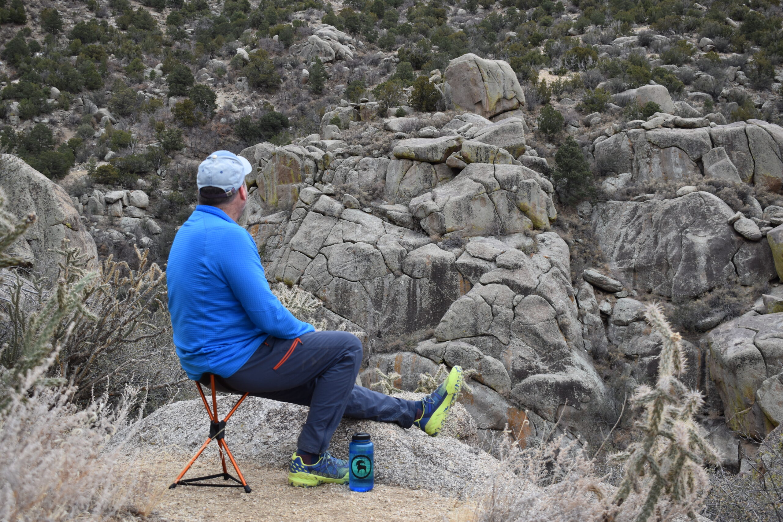 a backpacker rests alongside a desert canyon trail while sitting on the REI Stool with one leg extended and one foot on the ground for balance and stability