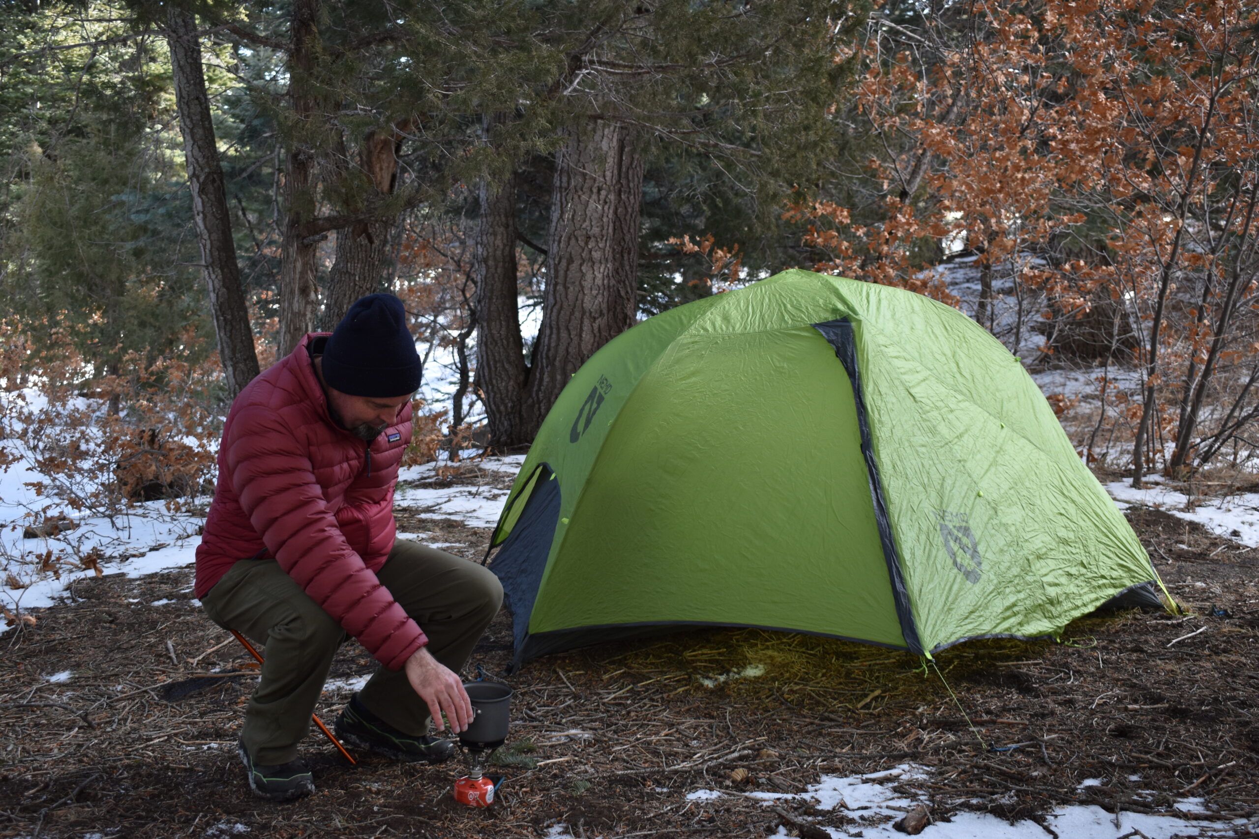 a camper sits on the stool as he leans forward to work with his camp stove outside of his tent