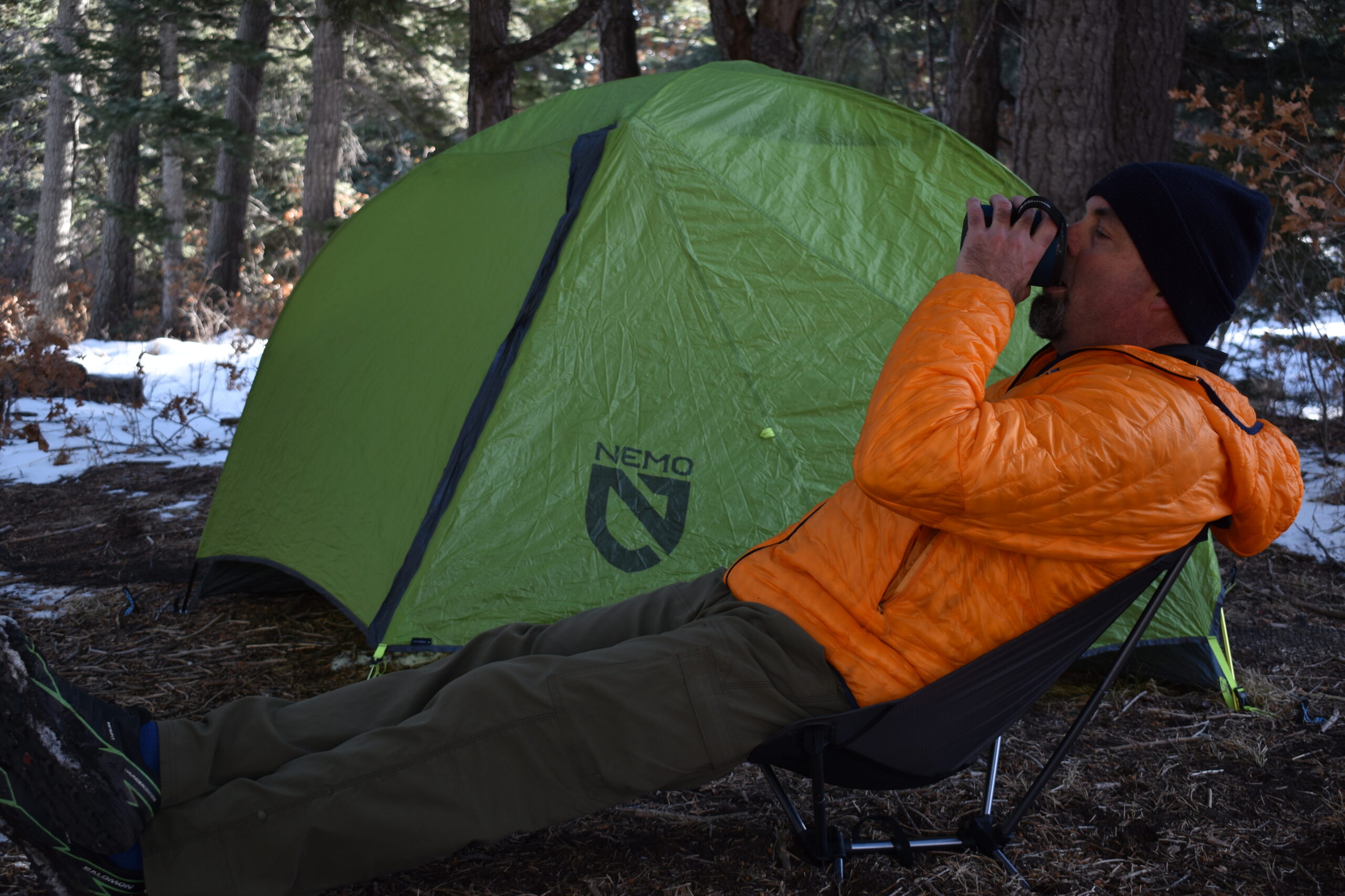 hiker in an orange puffy jacket sits on a backpacking chair in front of a green tent at a snowy campsite