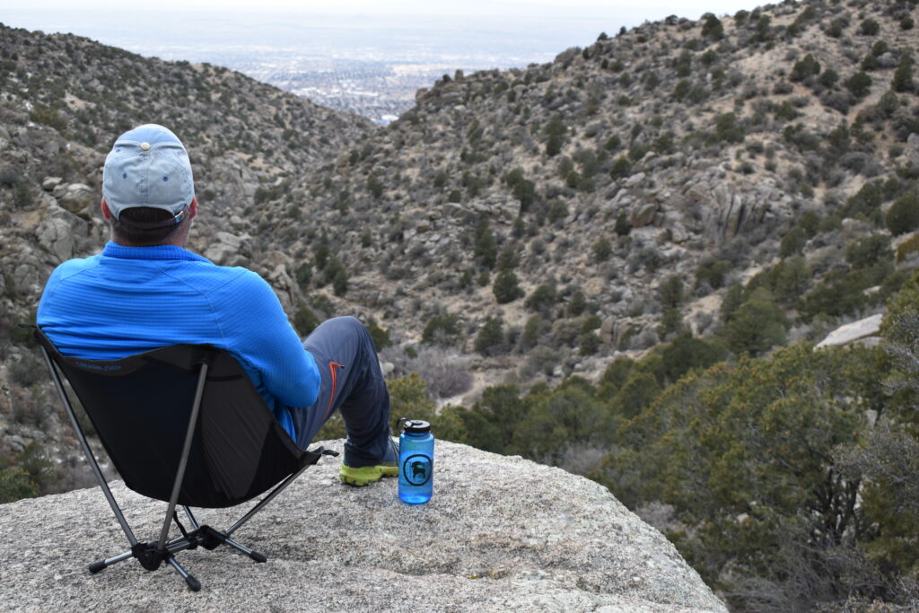 hiker sits on a rocky cliff overlooking a desert canyon while sitting on a camp chair