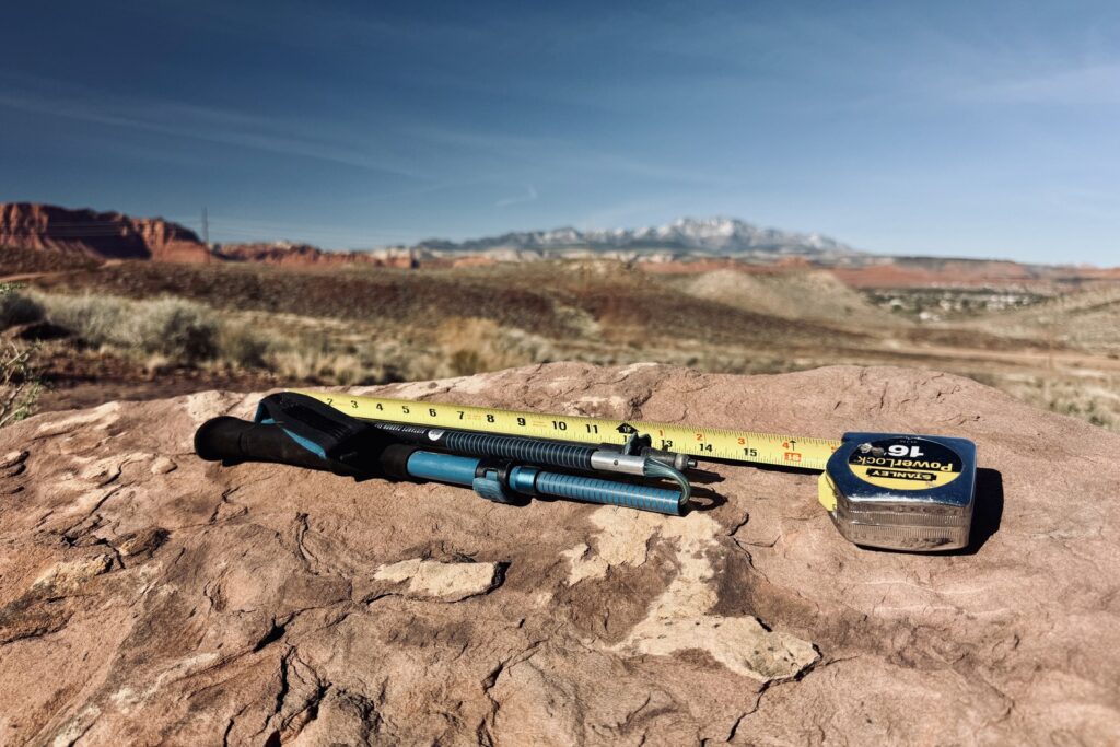 A close up of the Distance Carbon trekking poles laying next to an extended measuring tape on a red rock. There is red rock desert and a snowy mountain in the background.