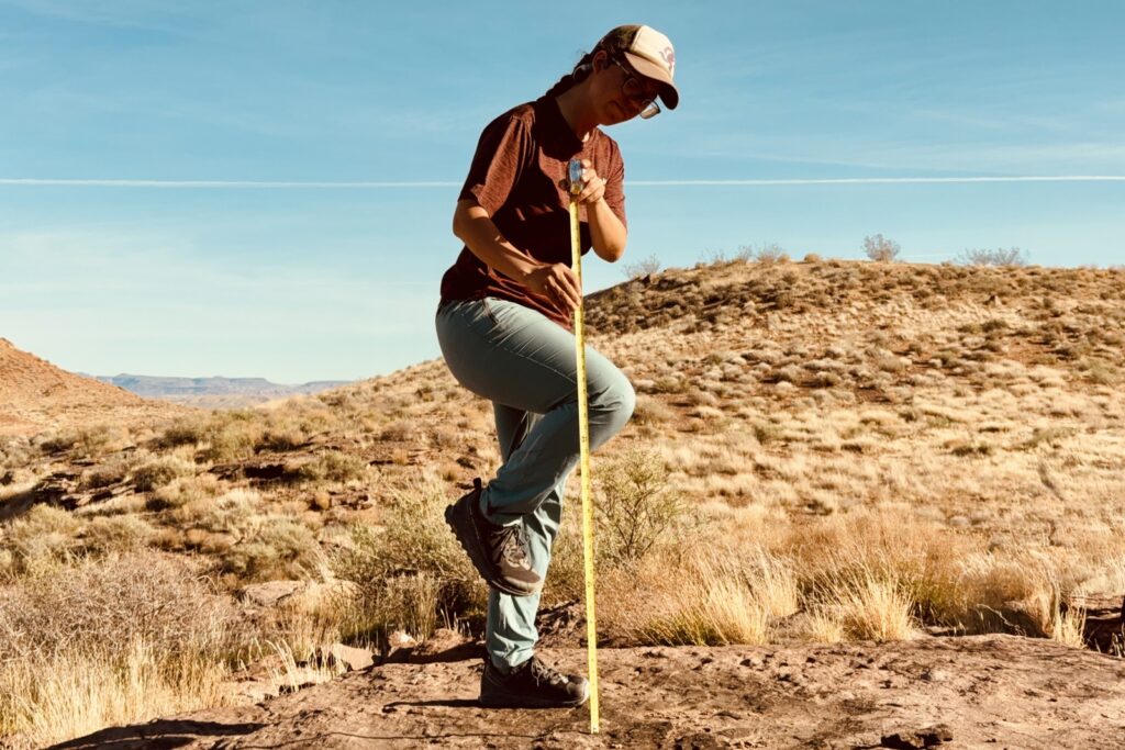 A person wearing the Baleaf pants is standing in a grassy, desert area. They are holding a measuring tape that is extended towards the ground next to their leg which is lifted up with the knee bent.