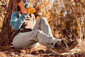 A person wearing the Baleaf pants is resting against a tree in the woods drinking water from a water bottle.