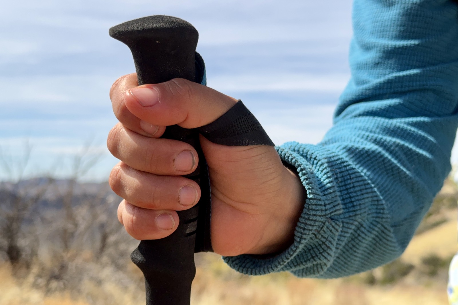 A close up of a person's hand holding the Distance Carbon trekking poles. The background is grassy with desert shrubs.
