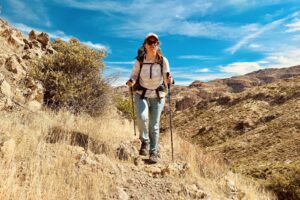 A person wearing a backpack is walking towards the camera on a trail using the Distance Carbon trekking poles. The area is rocky, grassy, and hilly.