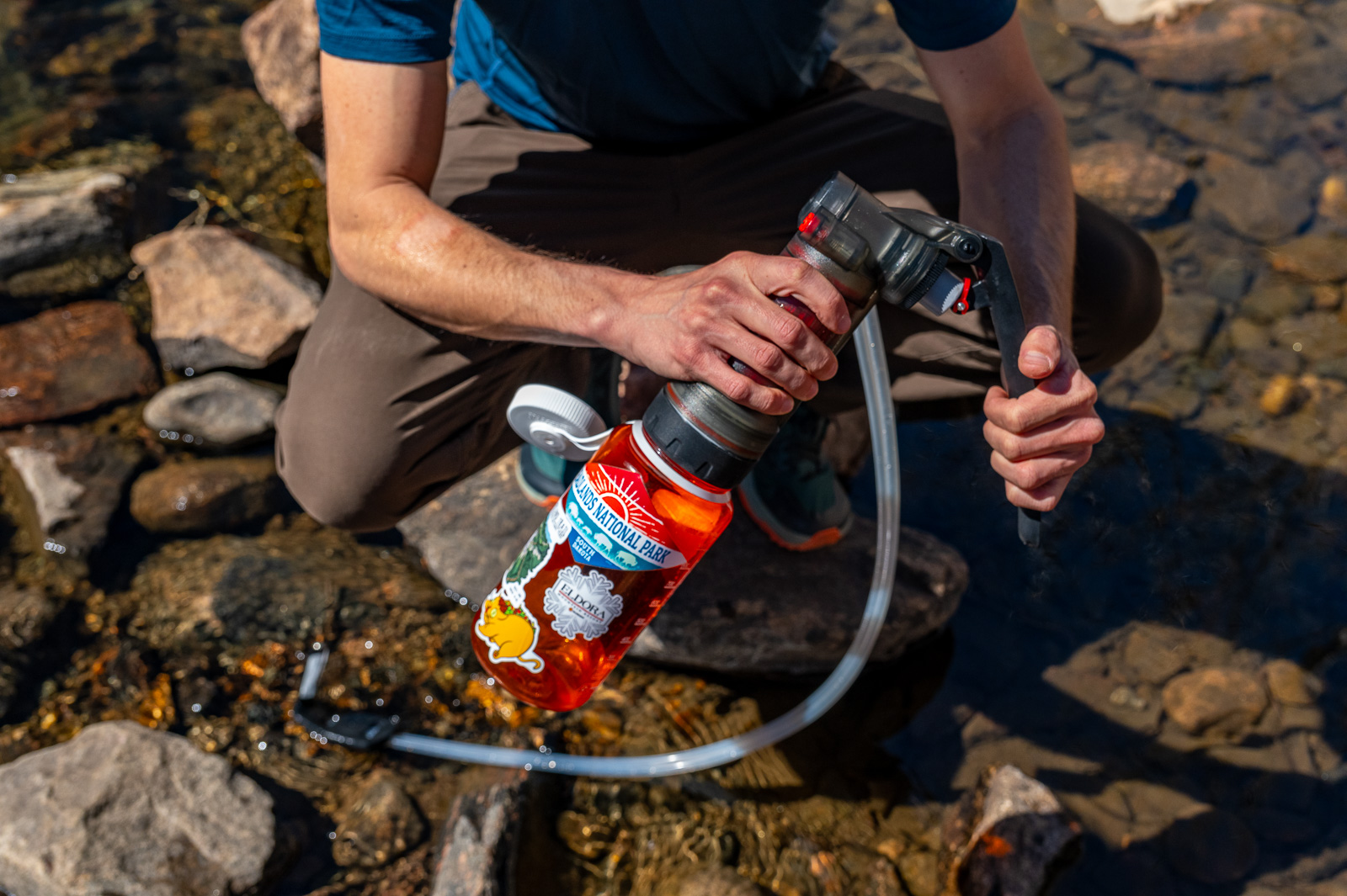 a hiker stoops on a rock in a creek while pumping water with the msr guardian and a nalgene bottle.