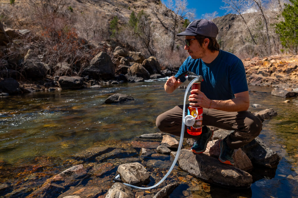 a hiker stoops on a rock in a creek while pumping water with the msr guardian and a nalgene bottle.