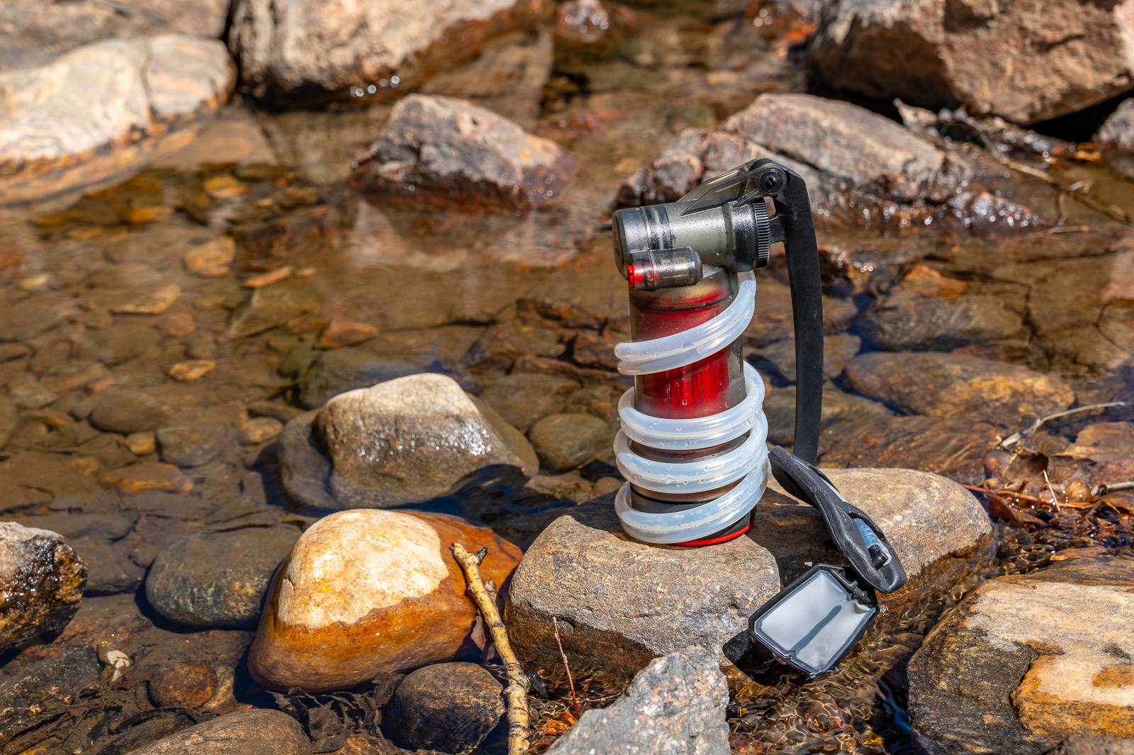 A pump water filter sits on a rock in a creek with the hose wrapped around it