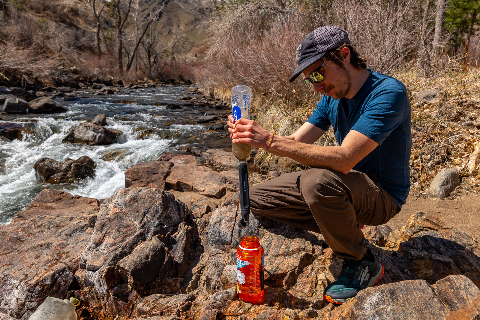 A hiker sits by a creek and squeezes water through the peak straw into a nalgene bottle.
