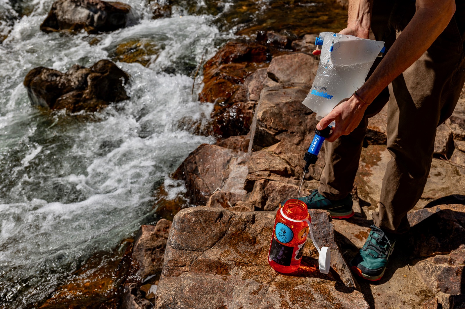 A hiker filters water with a bladder and the sawyer mini next to a creek