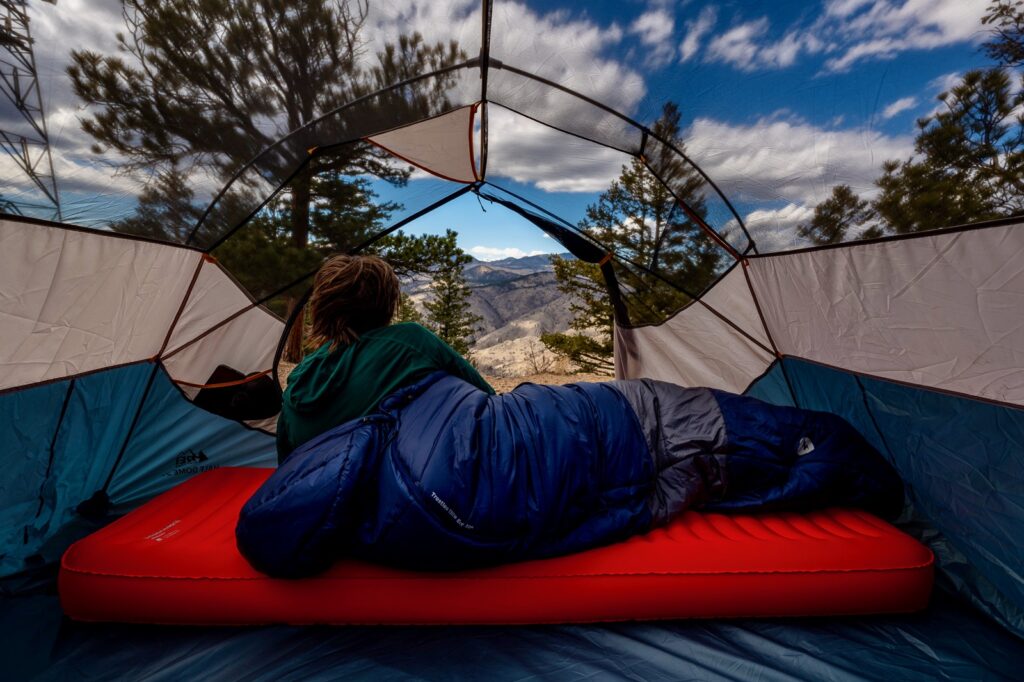 A hiker lays in a tent on a sleeping pad and sleeping bag looking out at a mountain view.