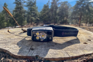 A black headlamp sits on a stup in a forest of pine trees. The sun is out.