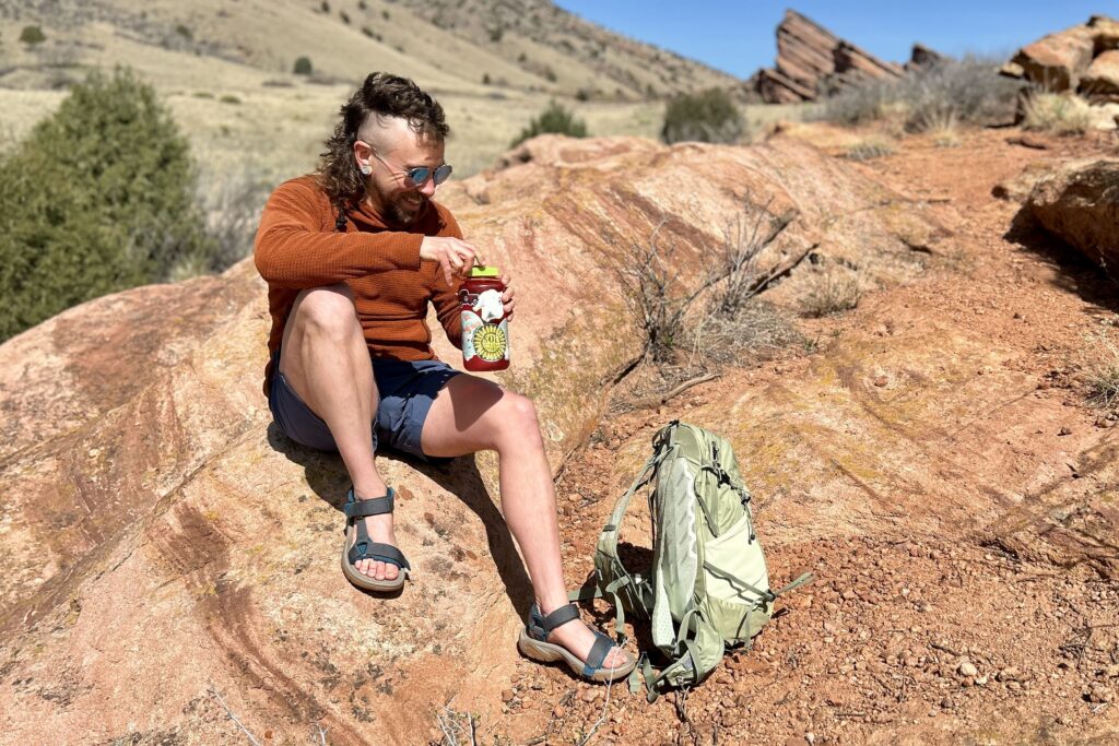 A man smiles as he sits on a rock in a desert setting and opens a water bottle with a backpack at his feet.