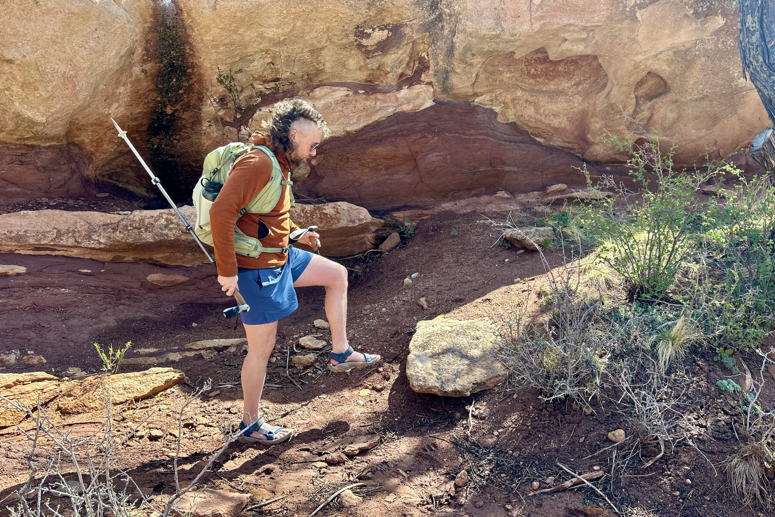 A man wearing a backpack and shorts and hiking sandals walks through a desert setting