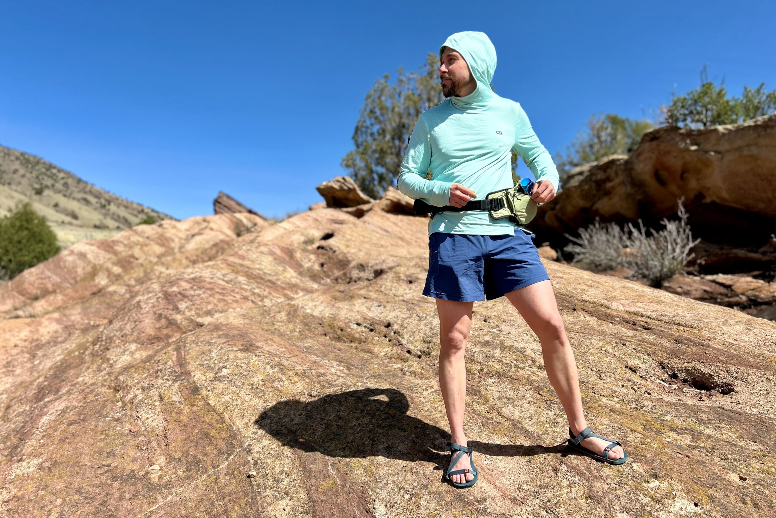 A man in a sun hoodie looks off into the distance on a sunny day while standing on some red rocks