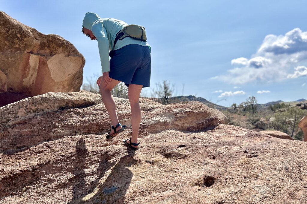 A man wearing a sun hoodie, shorts and sandals bends over as he climbs up a steep rock on a sunny day.