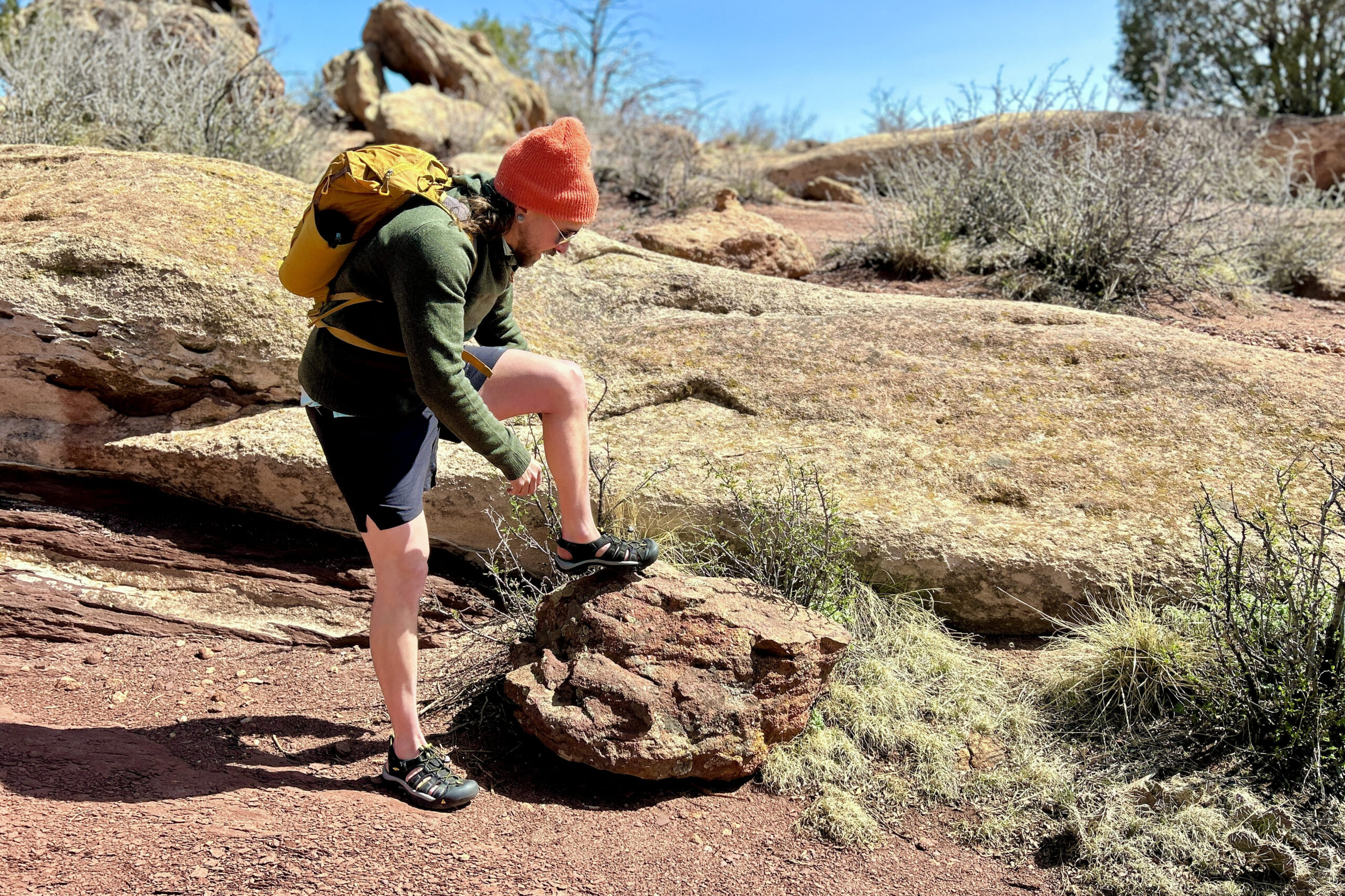 A man wearing a backpack and a hoodie leans over to adjust ahis sandal while stepping up on a rock on a sunny day.