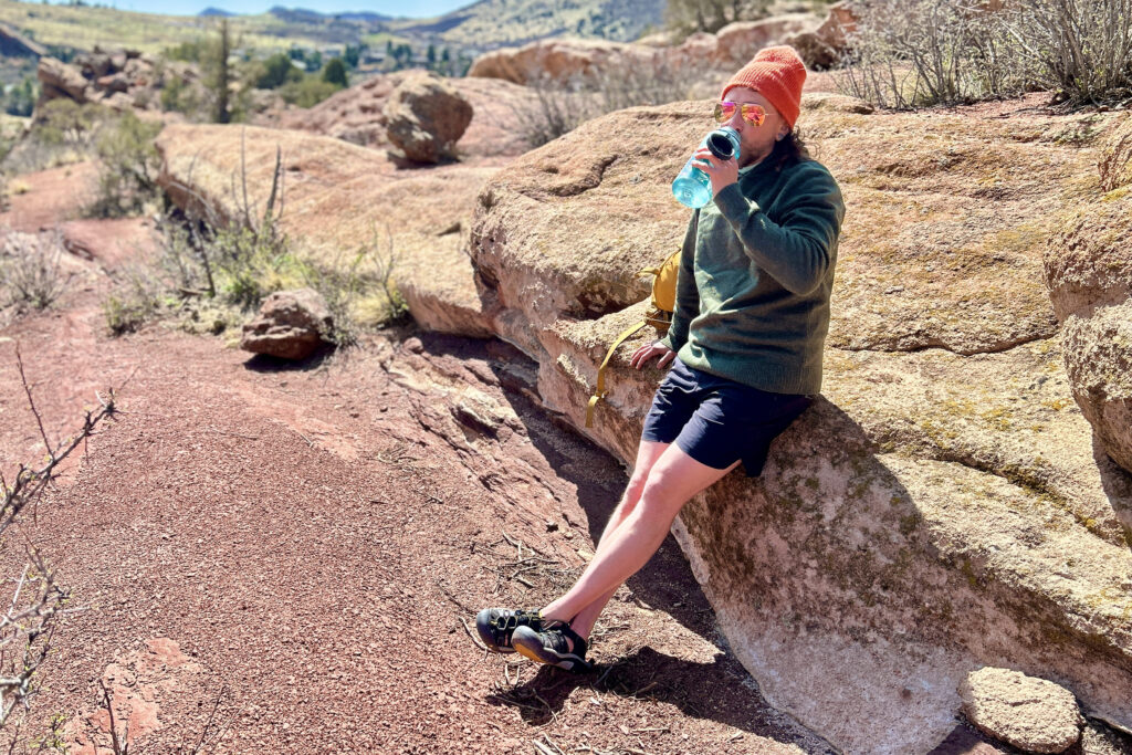 A man in a hoodie and warm hat leans on a big rock in a desert setting.