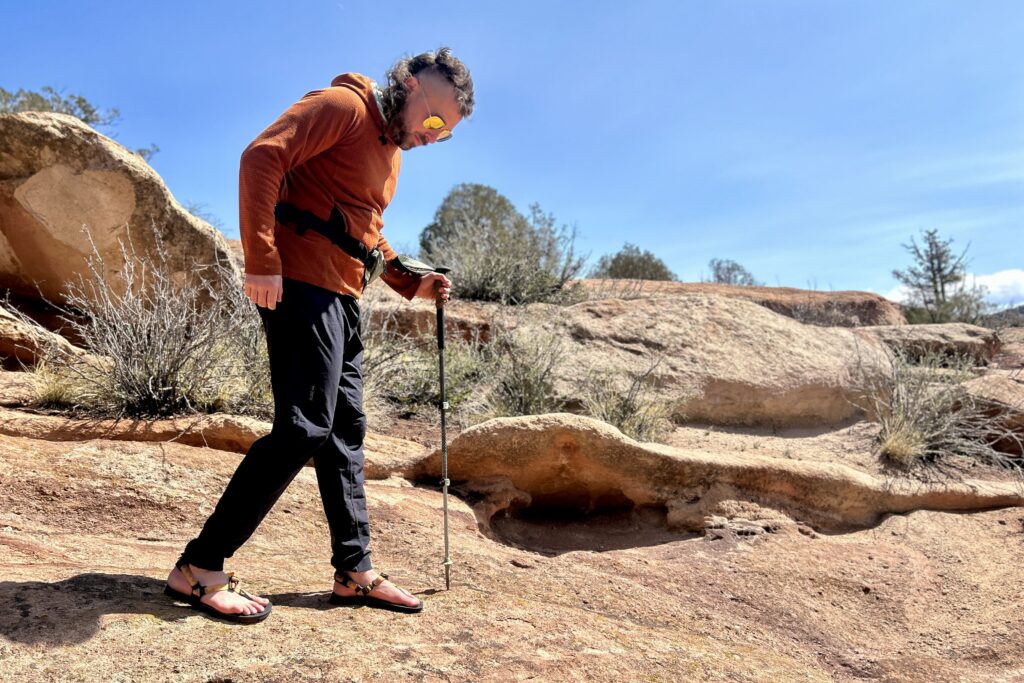A man walks through a desert setting holding onto a hiking pole, looking down at his feet.