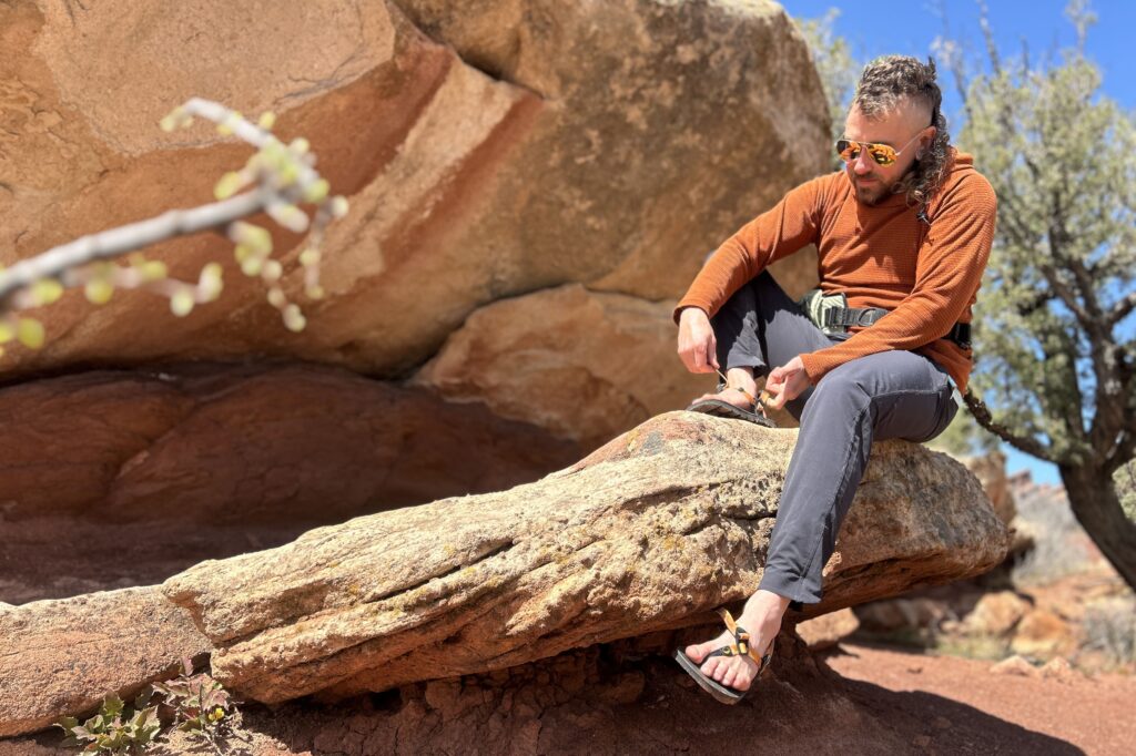 A man in a hoodie and pants sitting on a rock putting on a pair of sandals