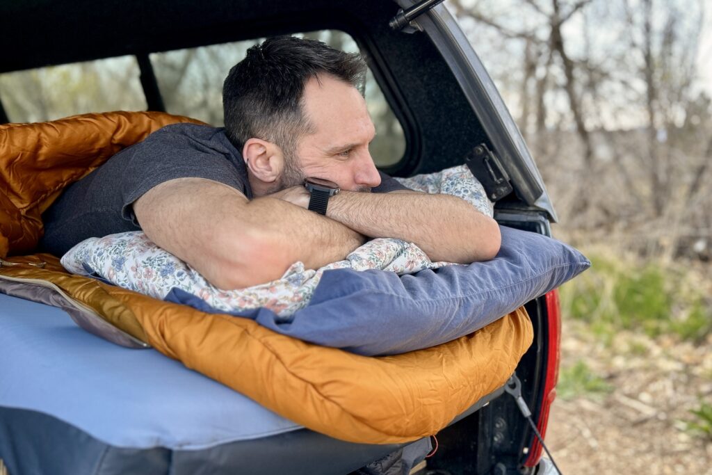 Close up image of a man resting on two pillows in a sleeping bag on a mattress in the back of a pickup truck.
