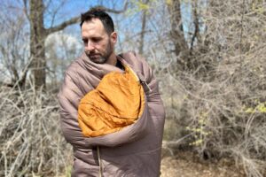 A man from the waist up is wrapped in a sleeping bag, standing outside and looking down at the ground