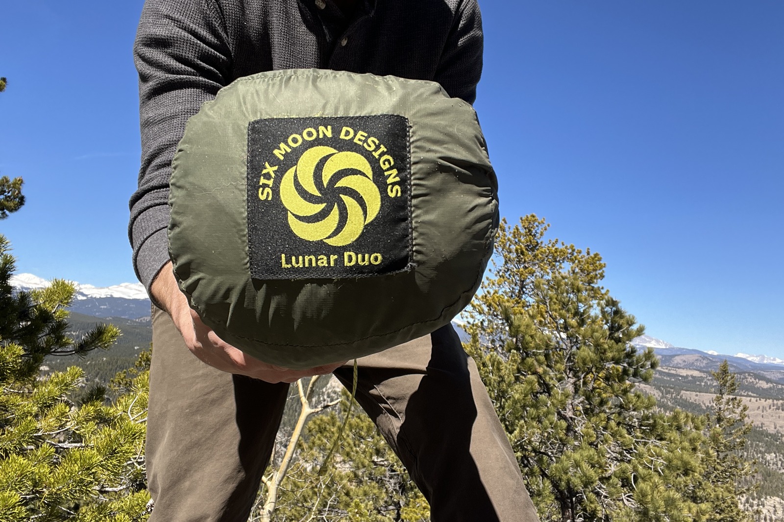 a hiker holds a tent in its stuff sack up to the camera with a logo patch reading "six moon designs - lunar duo"