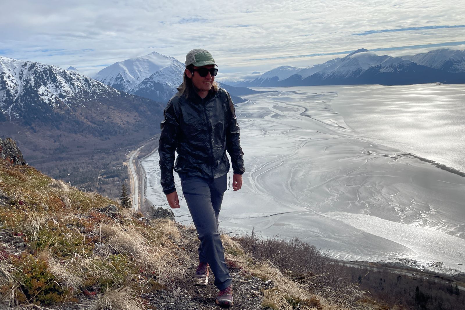 A man walks along a mountain trail with the ocean in the background.