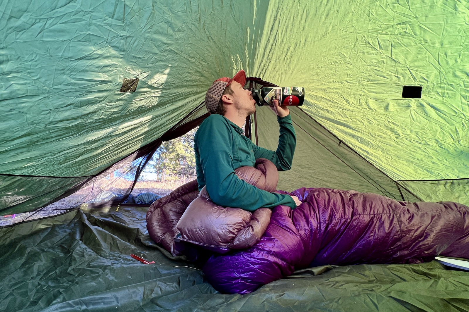 a hiker sits in a green tent and sips from his water bottle