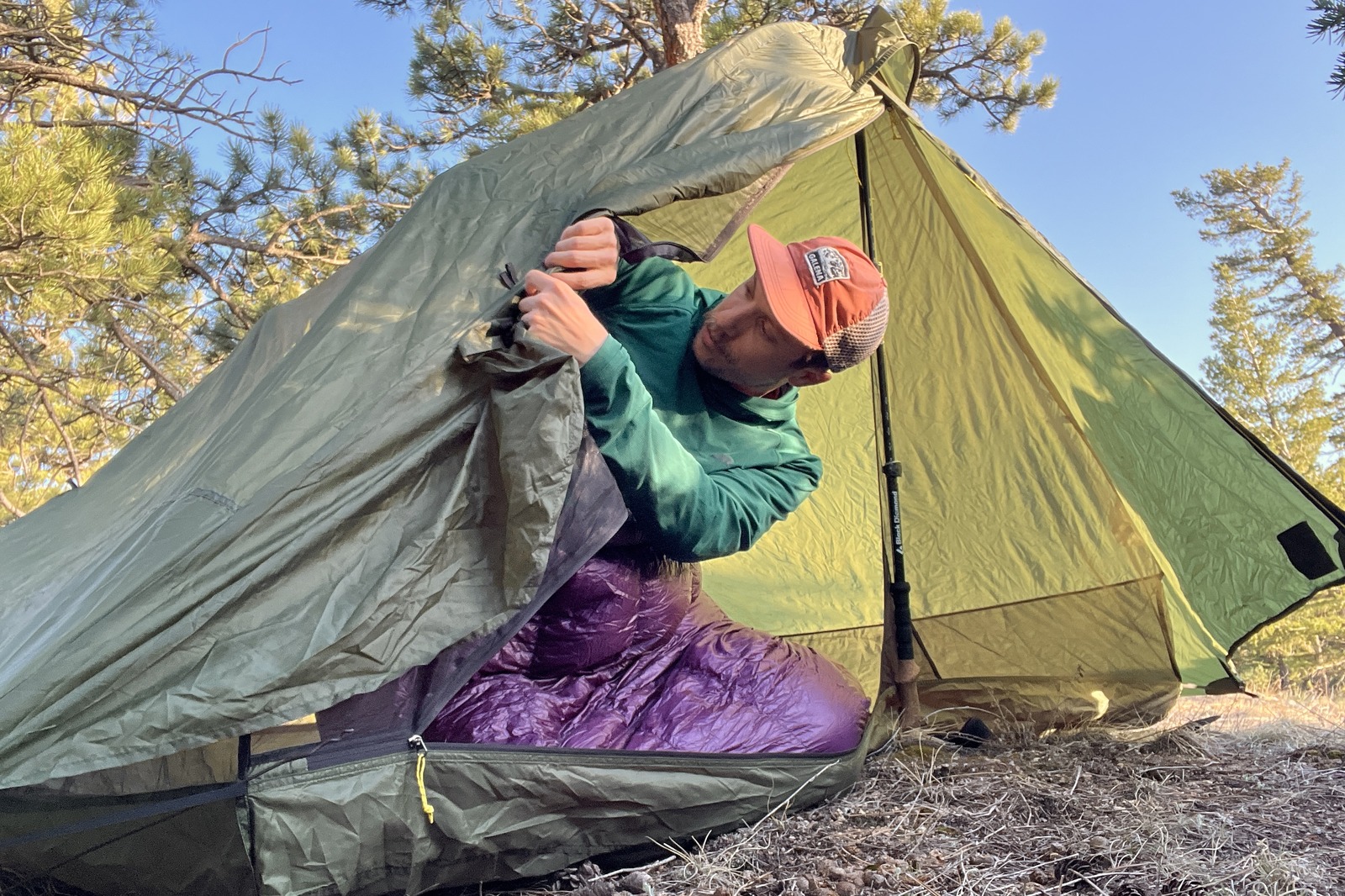 a hiker leans out of a green trekking pole tent to tie back the door