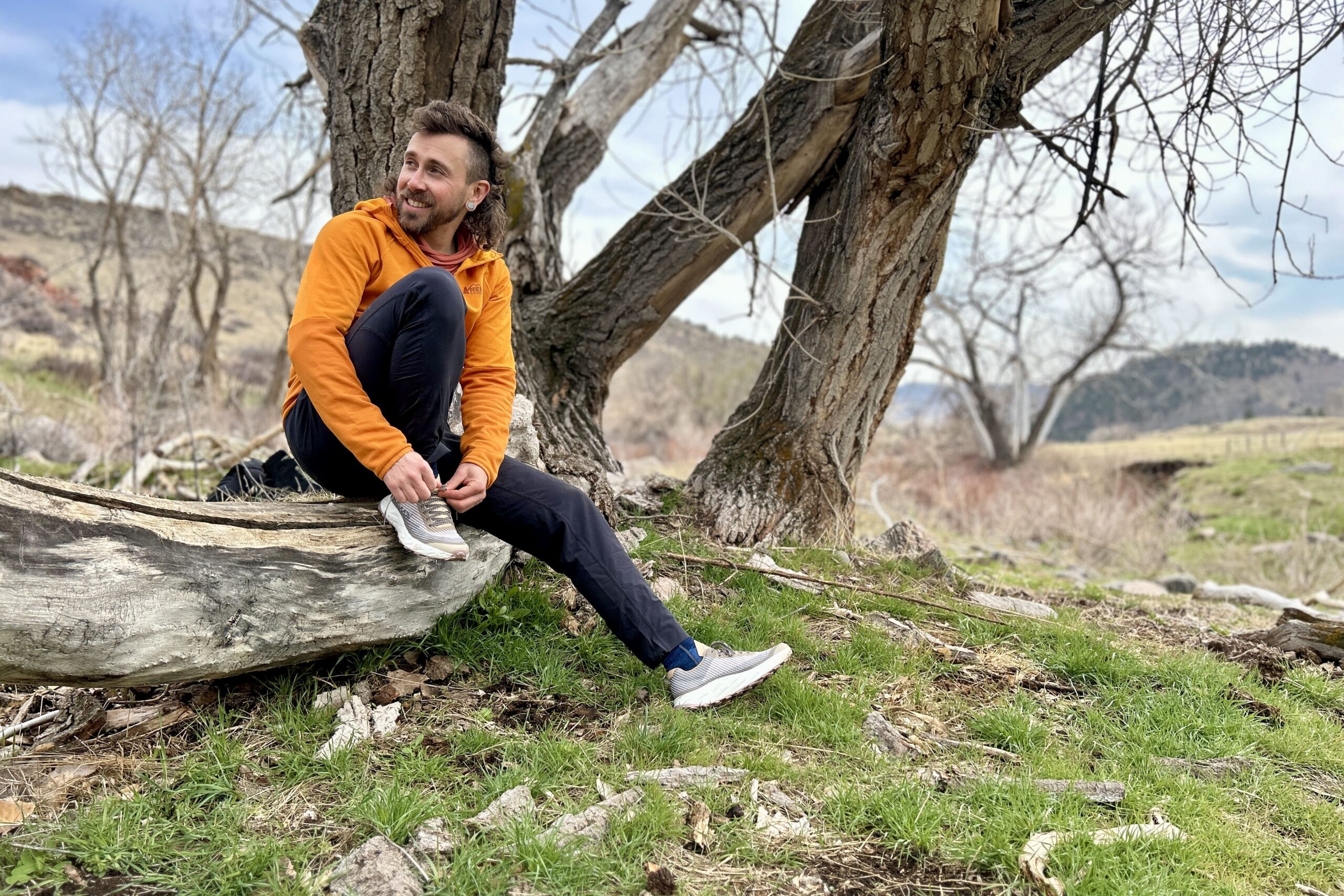 A man sits on a log smiling and looking off in the distance as he ties his trail running shoes.