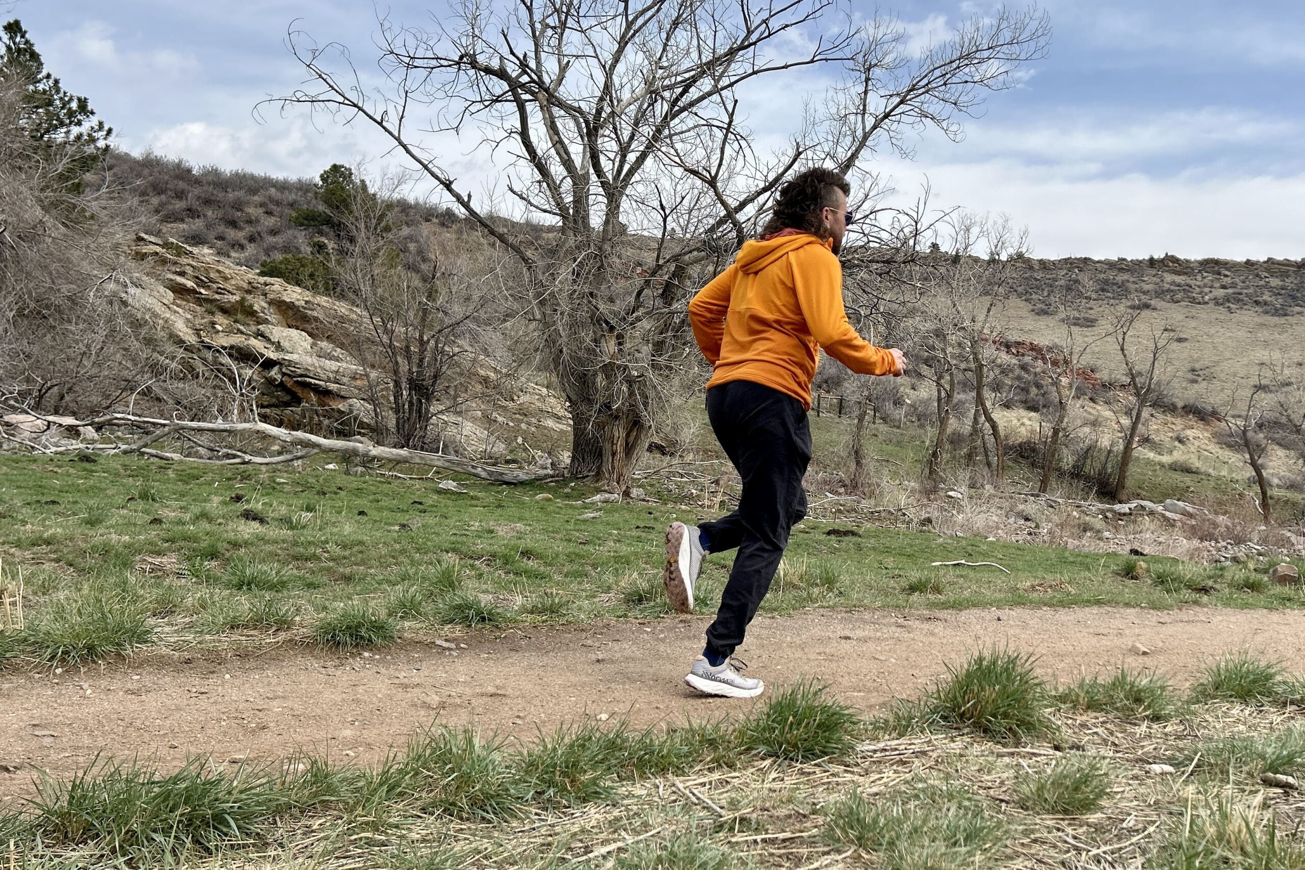 A man in an orange hoodie and black pants jogs down a trail in a rocky mountain setting.
