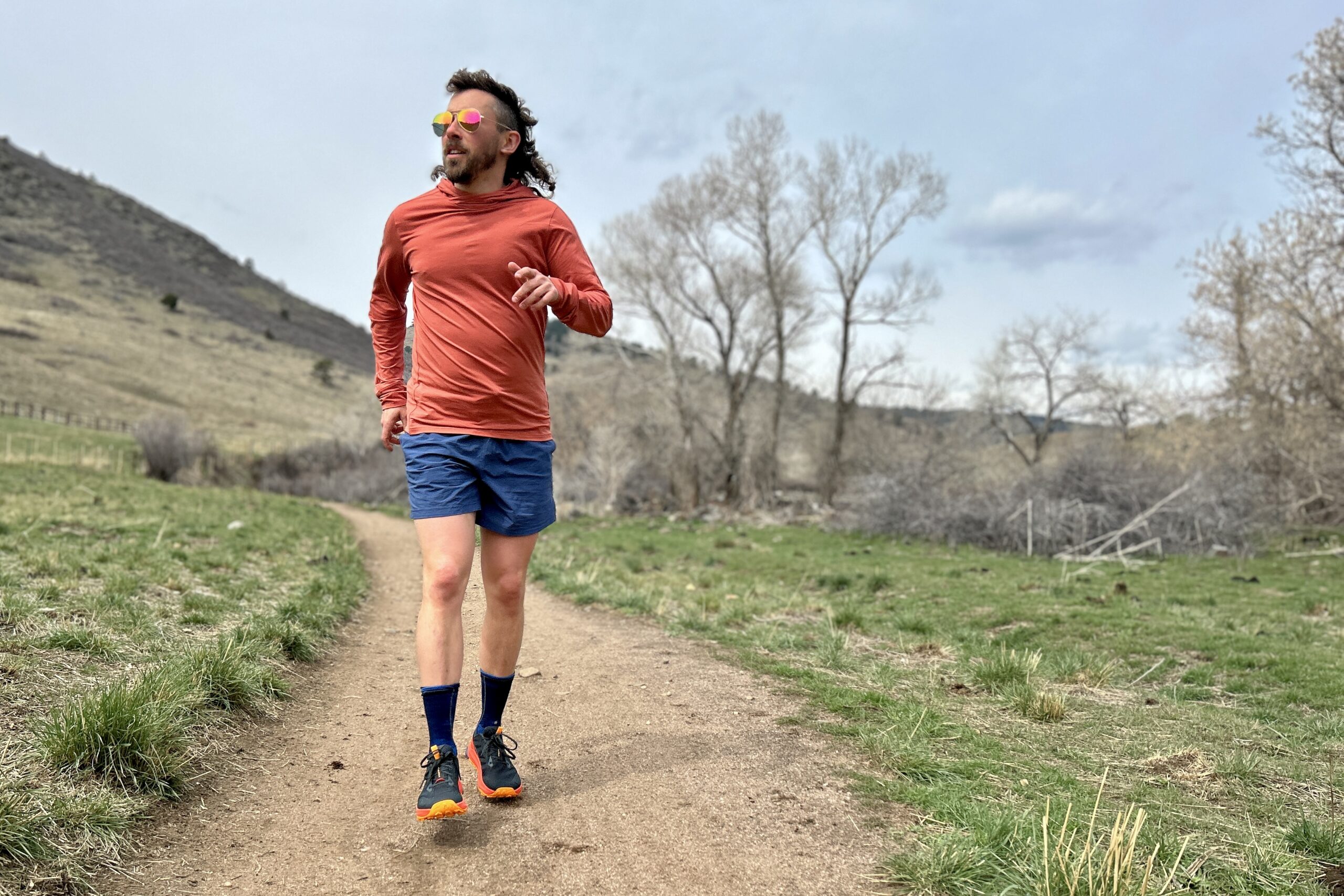 A man running down a trail towards the camera wears sunglasses and trail running attire in a hilly landscape.