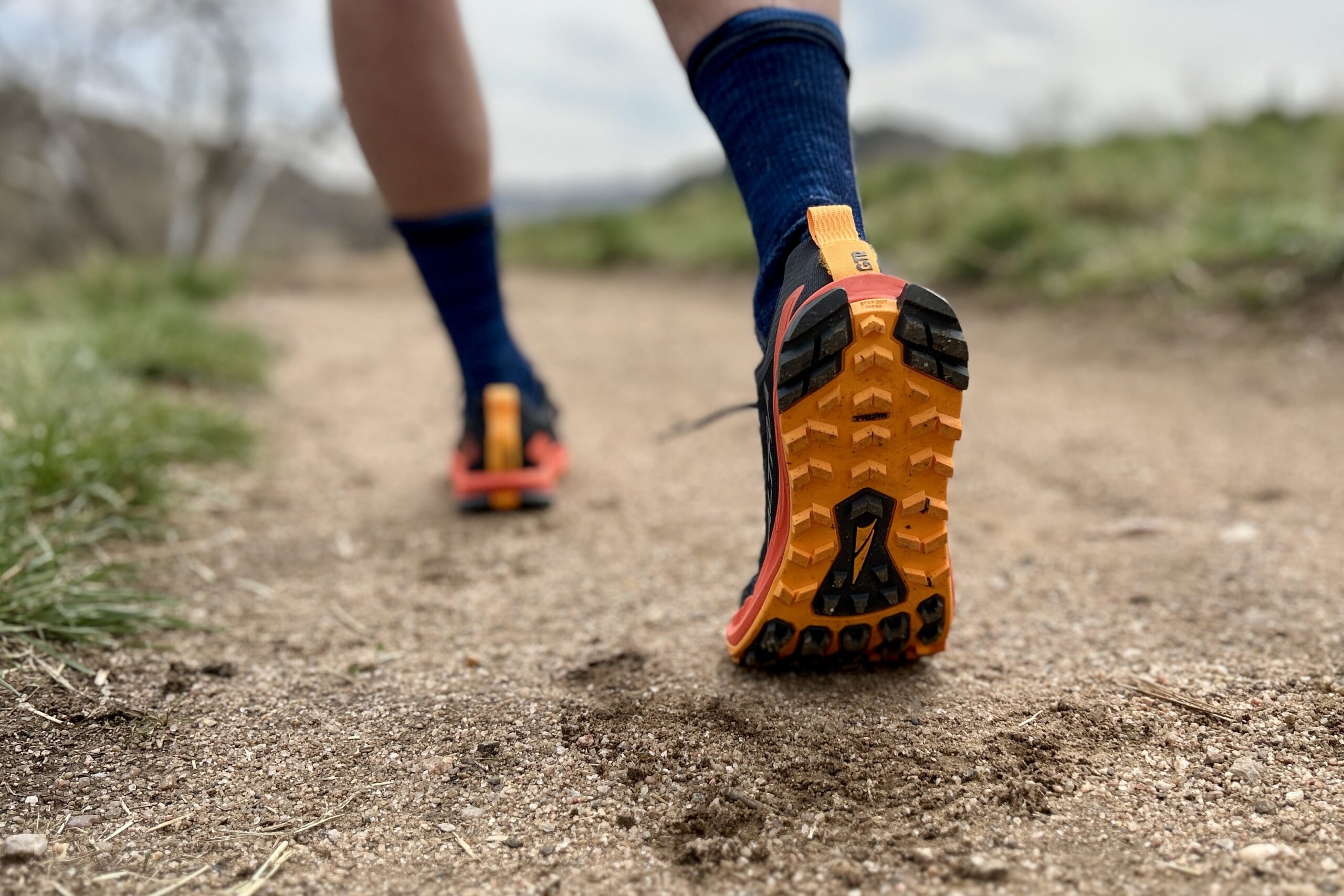 Close up from behind of a person wearing trail running sheos walking down a trail. the lug pattern is visible on one shoe.