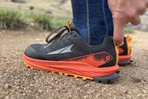 A person's finger is looped through the heel loop on a trail running shoe. Image is close up and centered on the shoe.